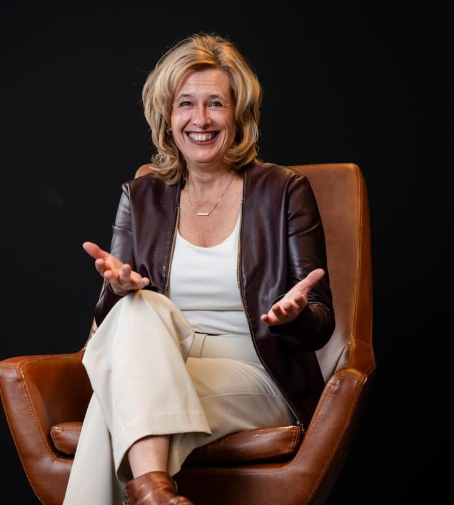 Portret van teamcoach en organisatiecoach Ine van den Heijkant in een lederen stoel lachend met de handenpalmen op